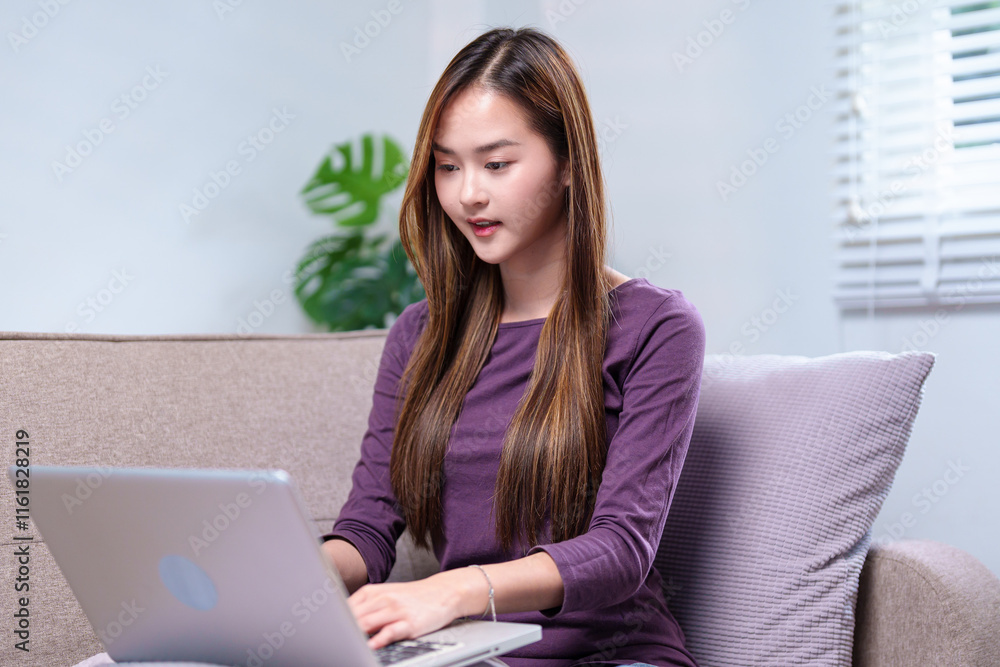 A young woman working on a laptop while sitting on a sofa, smiling and appearing focused, in a cozy home setting A young woman working on a laptop while sitting on a sofa, smiling and appearing focused, in a cozy home setting