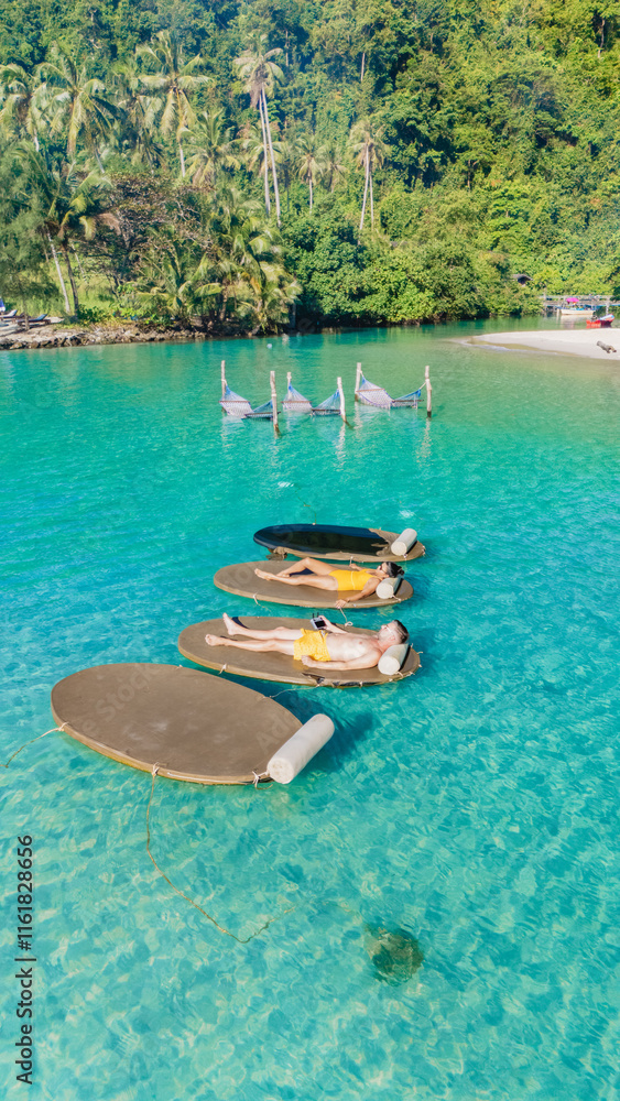 Obraz premium Under the warm sun, people enjoy leisure time on floating loungers amidst the clear turquoise waters of Koh Kood Thailand. Surrounded by lush greenery, the tranquil atmosphere invites relaxation.