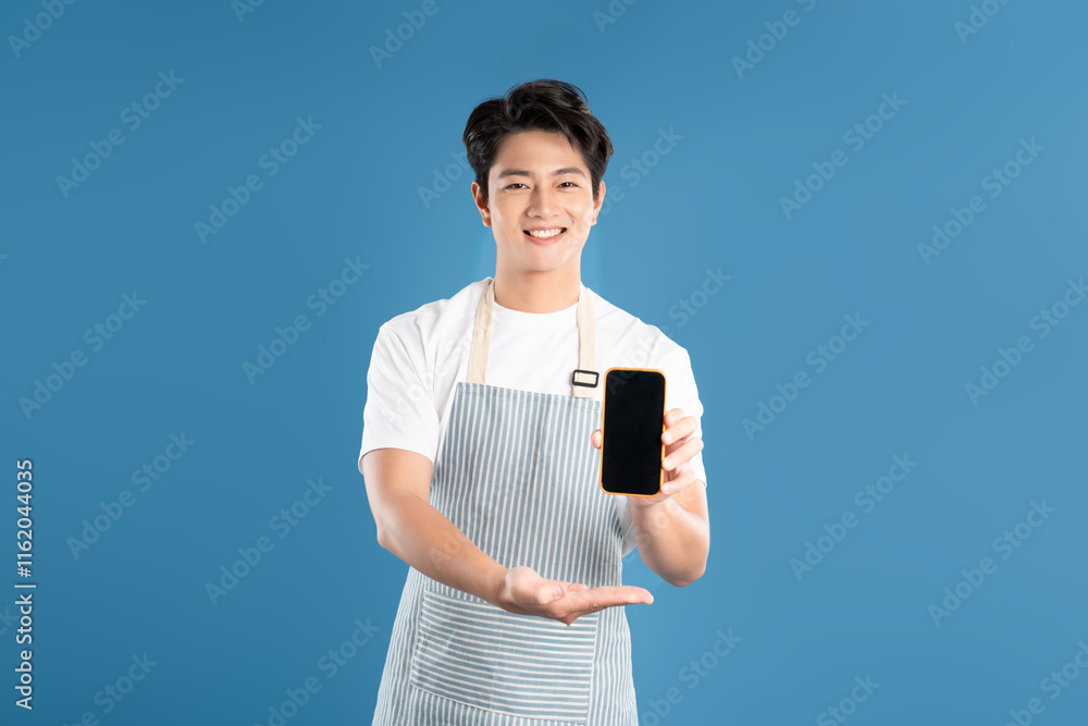 Portrait of young asian man wearing apron and using phone on blue background Portrait of young asian man wearing apron and using phone on blue background