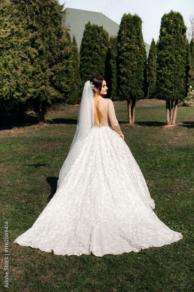 woman in a wedding dress standing on the grass against the backdrop of tall trees. White dress with a long train and open back. woman in a wedding dress standing on the grass against the backdrop of tall trees. White dress with a long train and open back.