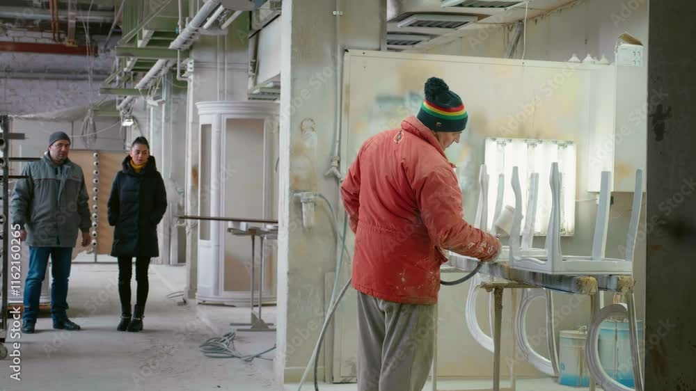 Man in a carpentry shop spraying paint on upside-down chair frames.
