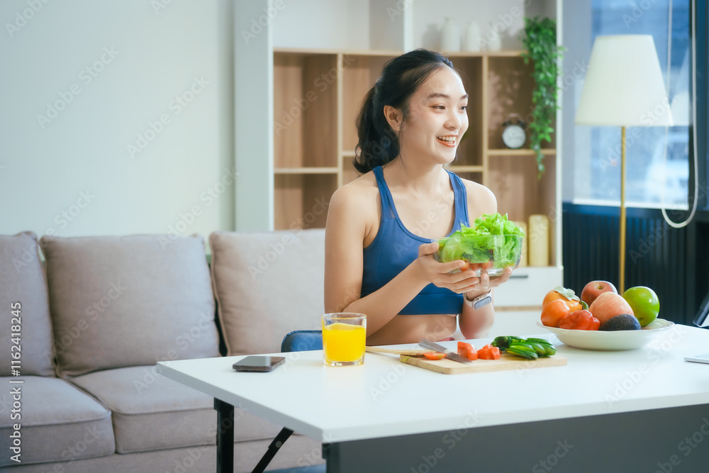 A happy Asian woman in sportswear sits on a sofa at a table, enjoying a salad bowl, fruits, fresh vegetables, orange juice, and energy supplements after a home fitness workout A happy Asian woman in sportswear sits on a sofa at a table, enjoying a salad bowl, fruits, fresh vegetables, orange juice, and energy supplements after a home fitness workout
