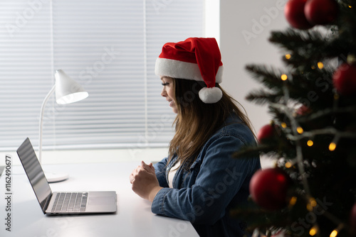 A woman wearing a festive Santa hat is engaged in an online activity at her holidaydecorated desk, surrounded by cheerful lights and ornaments, capturing the essence of the joyful winter season photo