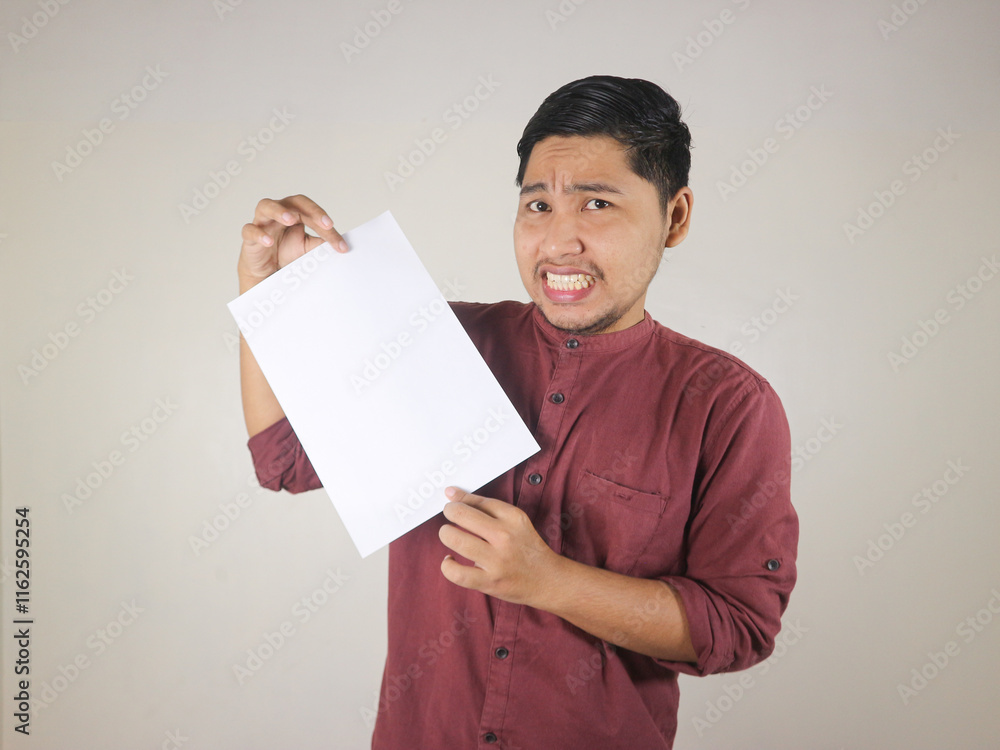 Asian man in red shirt holding white paper with worried and disappointed expression Asian man in red shirt holding white paper with worried and disappointed expression