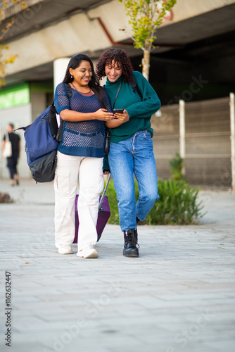 Two women walking with luggage and checking mobile phone on a city sidewalk photo