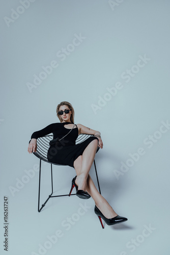 Attractive caucasian woman wearing black dress and sunglasses sitting on white studio background