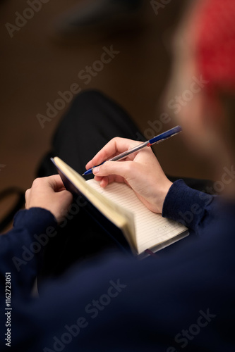 Person is writing with pen in notebook with his right hand indoors. Student in red hat is outlining information and taking notes in notebook. Planning in journal. Study. Exam. Training courses.