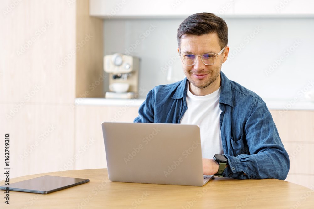 Young man working on laptop at kitchen table while enjoying remote work arrangement Young man working on laptop at kitchen table while enjoying remote work arrangement