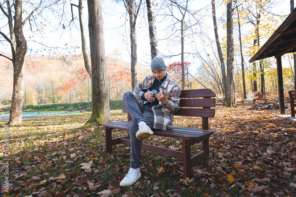 Fototapeta premium Enthusiast sits on a wooden bench in a park, playing the ukulele amidst autumn leaves. Surrounded by trees and bathed in soft sunlight, he enjoys a tranquil moment filled with creativity