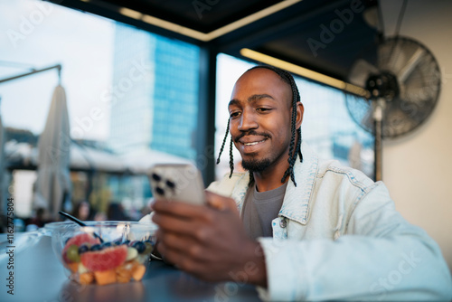 Smiling black man using smartphone and eating fruit salad at outdoor cafe