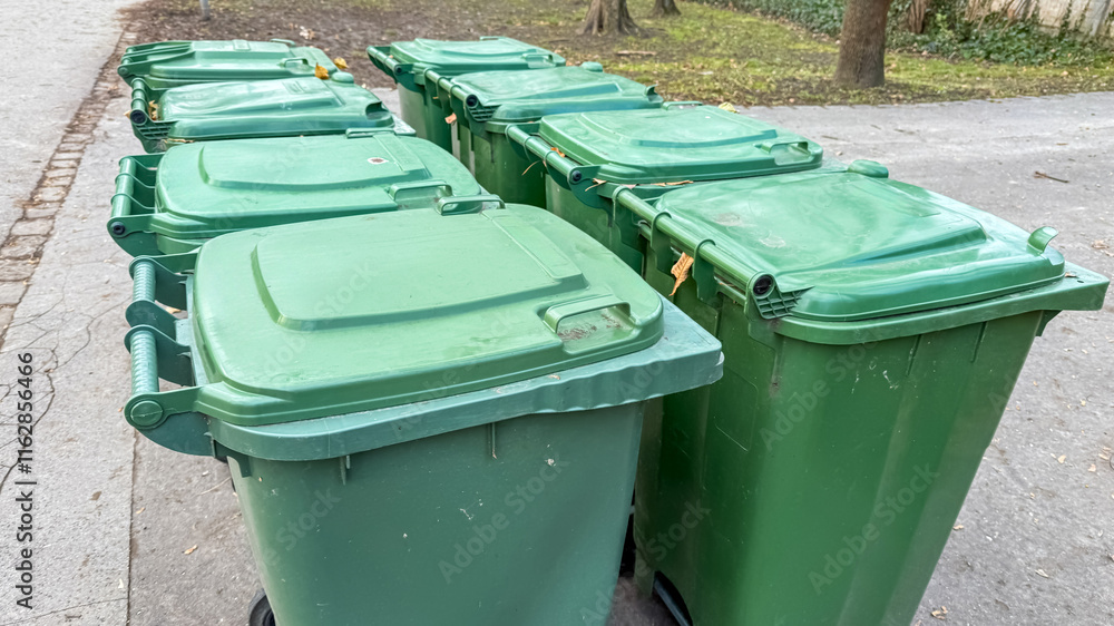 Multiple green trash bins lined up on a pavement highlight waste management and recycling issues in urban sustainability initiatives Multiple green trash bins lined up on a pavement highlight waste management and recycling issues in urban sustainability initiatives