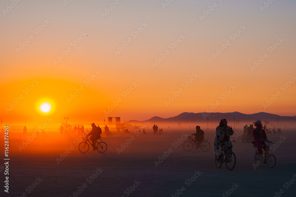 A warm orange sunset illuminates a desert festival scene with cyclists, art installations, and distant mountains in Black Rock Desert, Nevada.