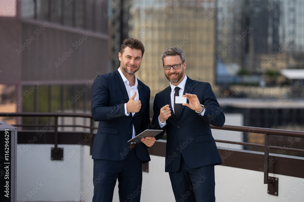 A man and a man exchanging a business card. one businessman gives another businessman his business card outside an office center. a man in a business suit holding a credit card A man and a man exchanging a business card. one businessman gives another businessman his business card outside an office center. a man in a business suit holding a credit card