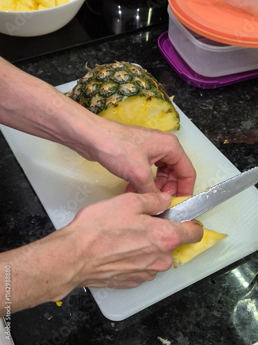 A person slicing a fresh pineapple on a white cutting board in a modern kitchen. The fruit is prepared for storage in containers, showcasing a healthy snack preparation process.