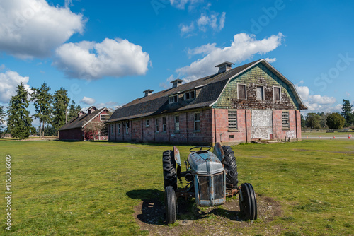 An old tractor sits in front of a large, weathered barn on a bright, clear day. Fluffy clouds scatter across the blue sky, and the lush green grass signifies an open, rural setting.