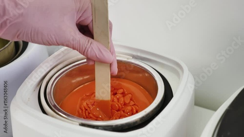 A depilatory master stirs orange granular wax into a wax melter with a wooden spatula, close-up photo