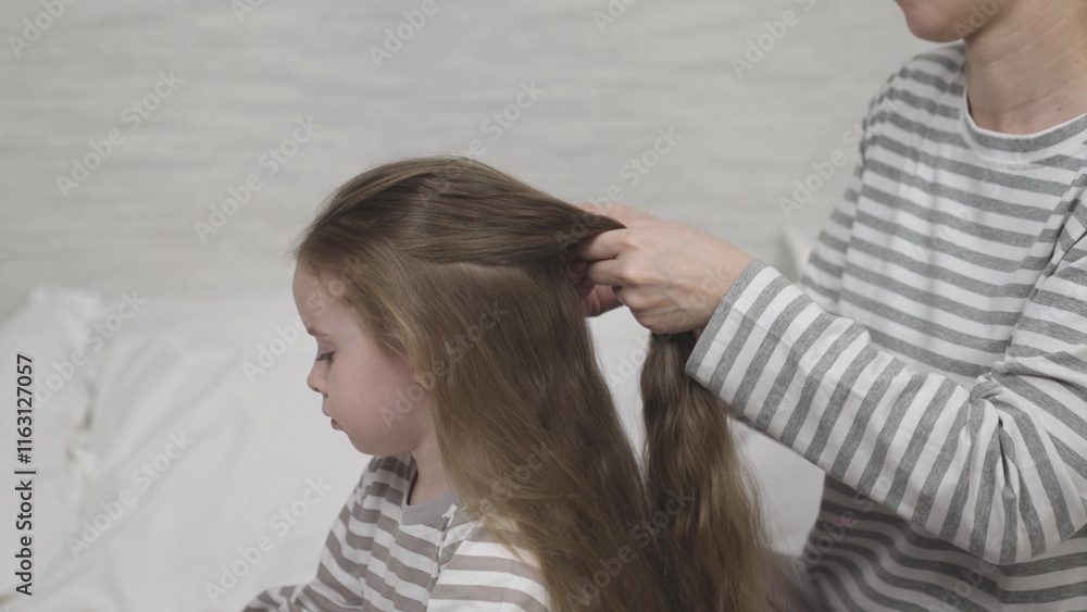 Fototapeta premium Mother braids child long brown hair. Mom creates hairstyle daughter brunette loose hair. Mum and preschooler girl take care hair closeup slow motion. Happy family. Mom kid together happy family indoor