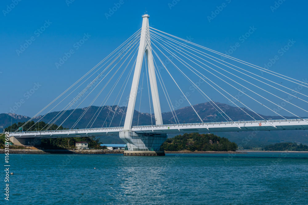 A cable-stayed bridge spanning across a ocean inlet with mountains in the background under a clear blue sky, located in Wando, South Korea A cable-stayed bridge spanning across a ocean inlet with mountains in the background under a clear blue sky, located in Wando, South Korea