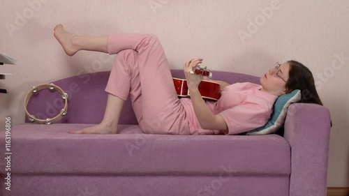 Young woman learning to play Spanish six string acoustic guitar while lying on her back on sofa at home. Side view of 18 year old model of Caucasian ethnicity. Female guitarist wears jeans, polo shirt photo