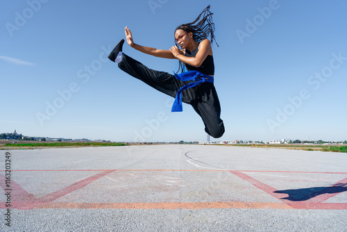 Chica experta en artes marciales haciendo un ataque saltando en el aire, en el paisaje infinito y minimalista de una pista de aterrizaje, un día soleado de verano con cielo azul. photo