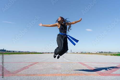 Chica luchadora de artes marciales saltando muy contenta con los brazos abiertos y una sonrisa una sonrisa, en el paisaje infinito y minimalista de una pista de aterrizaje un día soleado y cielo azul. photo