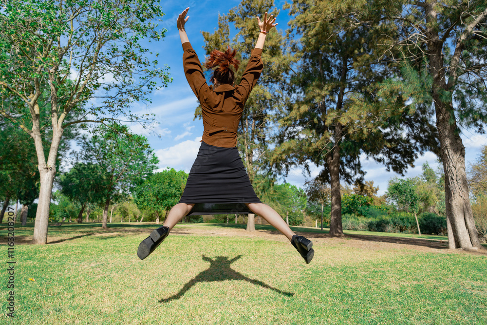 Mujer joven saltando muy alegre con los brazos abiertos en un parque un día soleado con cielo azul y nubes.