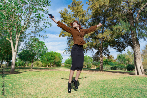 Mujer joven saltando muy alegre con los brazos abiertos en un parque un día soleado con cielo azul y nubes. photo