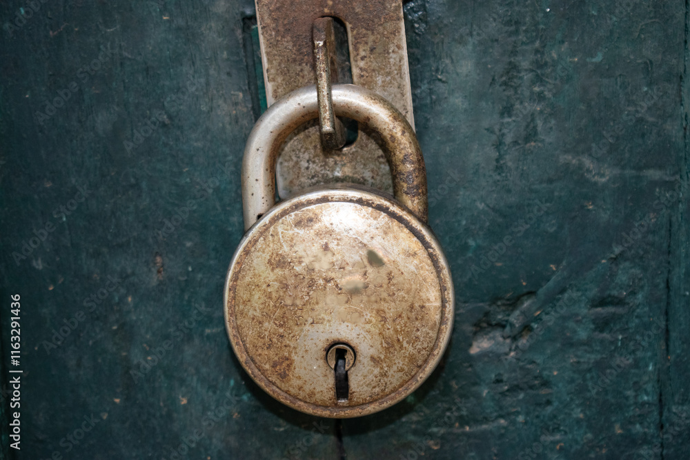 A close-up view of a circular, aged steel padlock secured to a latch on a weathered green wooden surface. The lock displays signs of wear and patina. A close-up view of a circular, aged steel padlock secured to a latch on a weathered green wooden surface. The lock displays signs of wear and patina.