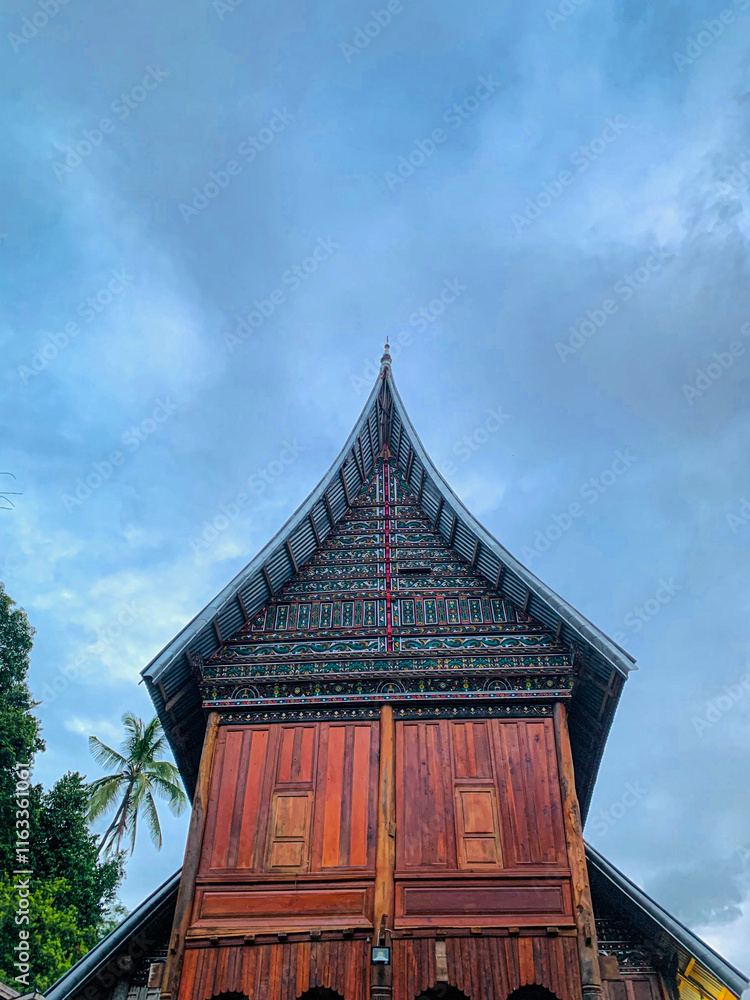 Detailed Roof Structure: A frontal view emphasizing the towering, curved rooflines of a Rumah Gadang, adorned with colorful, detailed motifs, standing against a serene, cloud-filled sky.