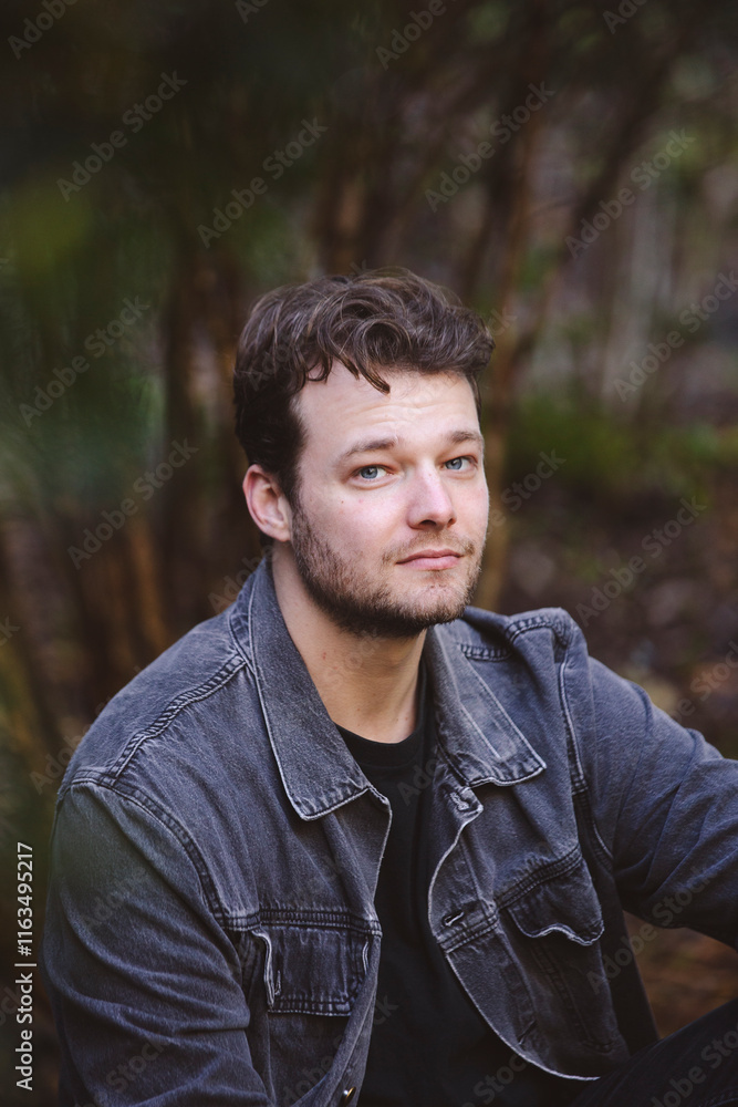 A young man in a dark denim jacket sits casually in a natural setting surrounded by lush greenery. His expression is calm and thoughtful as he gazes to the side, with soft light on his face A young man in a dark denim jacket sits casually in a natural setting surrounded by lush greenery. His expression is calm and thoughtful as he gazes to the side, with soft light on his face
