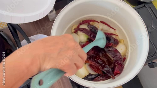 Hand stirs a mixture of dried chili and chopped onions, fresh ingredients in a pot. The setting reflects a home kitchen where traditional cooking techniques are being highlighted. photo