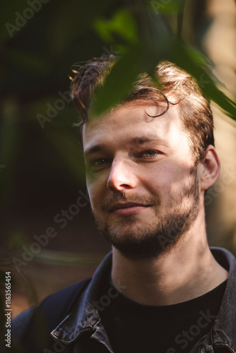 A candid portrait of a man partially hidden by green foliage, with sunlight softly illuminating his face. The natural background and shadowing create a serene and mysterious atmosphere