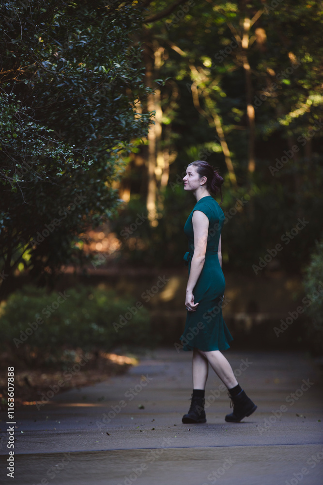 Obraz premium A woman walks confidently down a path in a lush green forest, wearing a dark green dress and black boots. Natural sunlight filters through the trees, creating a serene and vibrant atmosphere