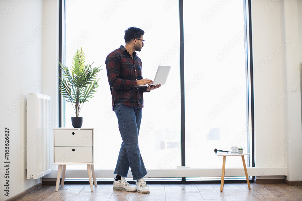Young Indian male standing by large window with laptop in modern office. Casual attire with flannel shirt and jeans. Bright and minimalist workspace with indoor plant and natural light. Young Indian male standing by large window with laptop in modern office. Casual attire with flannel shirt and jeans. Bright and minimalist workspace with indoor plant and natural light.