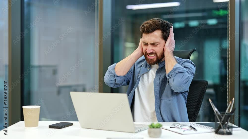 Frustrated businessman covering ears with hands while working on laptop in modern office. He is overwhelmed with information overload and stress. Frustrated businessman covering ears with hands while working on laptop in modern office. He is overwhelmed with information overload and stress.
