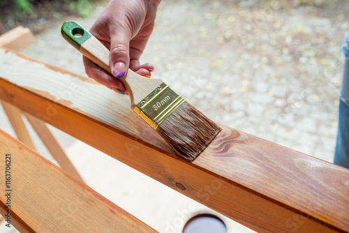 Close-up of a worker’s hand applying wood stain with a paintbrush on wooden beams during a DIY outdoor renovation or home improvement project