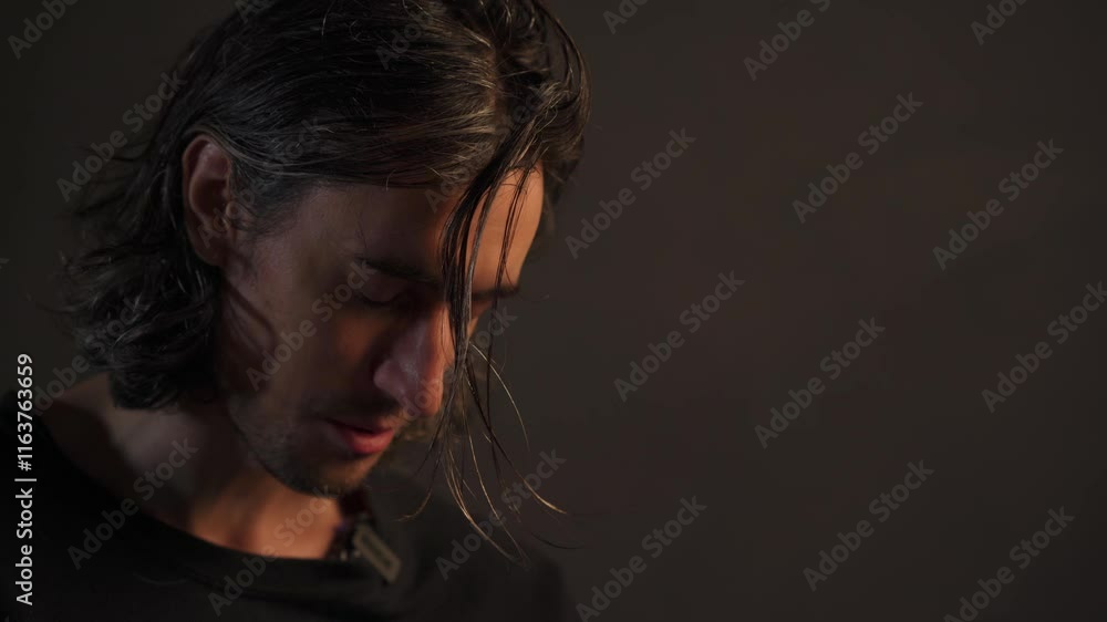 Mysterious Young Man Smiling A handsome European guy with long hair smiles softly in a dark atmospheric setting while speaking with a gentle demeanor. Mysterious Young Man Smiling A handsome European guy with long hair smiles softly in a dark atmospheric setting while speaking with a gentle demeanor.
