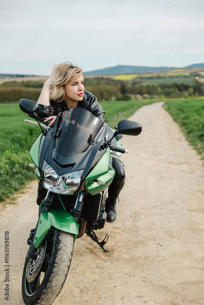 Obraz premium A stylish woman with red lipstick sits on a green motorcycle parked on a dirt road, wearing a black leather jacket. She gazes at the scenic view, evoking freedom and adventure.