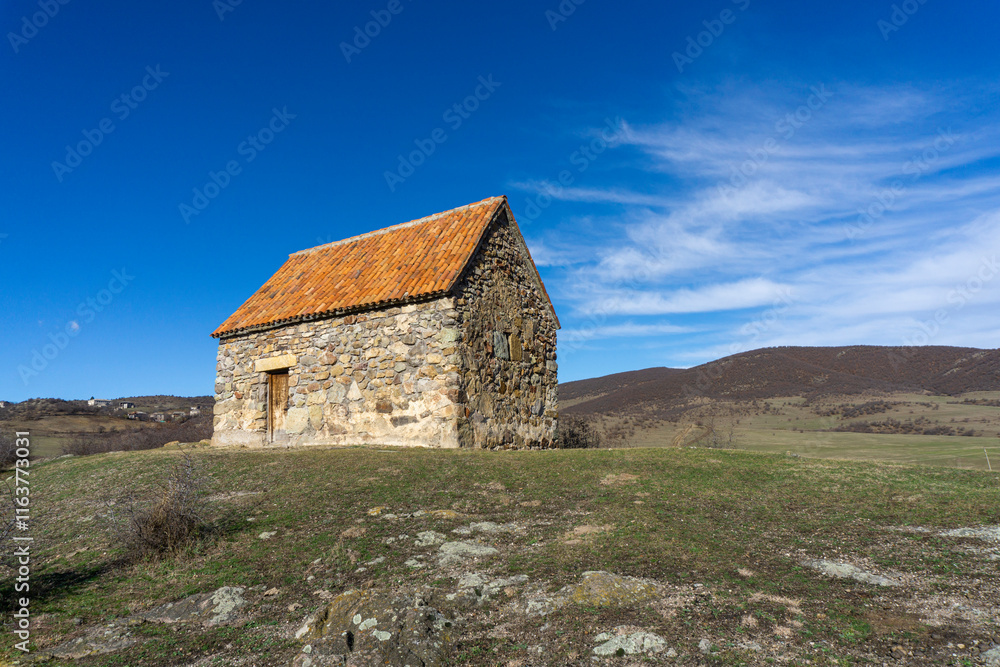 Fototapeta premium Small shurch on the rock. Wooden door. Orange tiled roof, stone walls. Green grass and bushes. Bright blue sly with clouds. Georgia