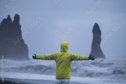 A person wearing a fluorescent yellow jacket and white waterproof pants standing with their back to the black sand beach in Iceland.