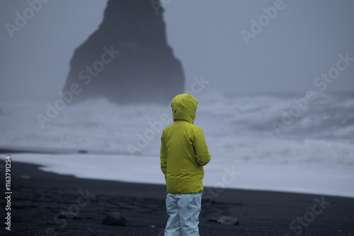 A person wearing a fluorescent yellow jacket and white waterproof pants standing with their back to the black sand beach in Iceland.