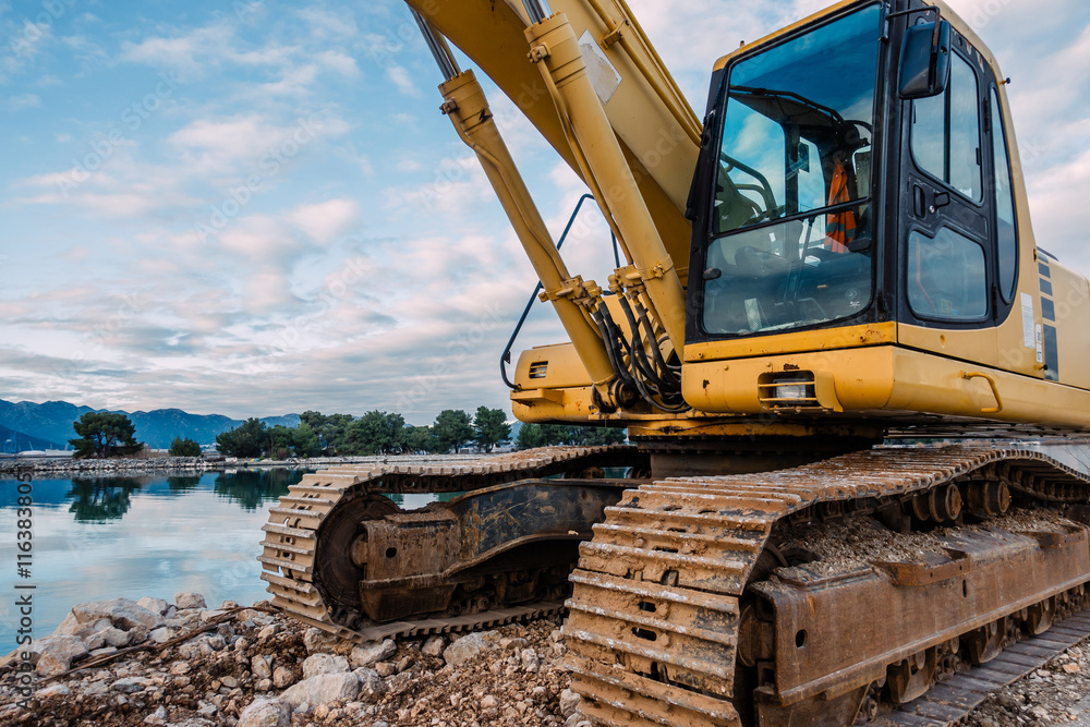 Heavy Yellow Excavator on Rocky Terrain at a Coastal Construction Site with Calm Water Heavy Yellow Excavator on Rocky Terrain at a Coastal Construction Site with Calm Water