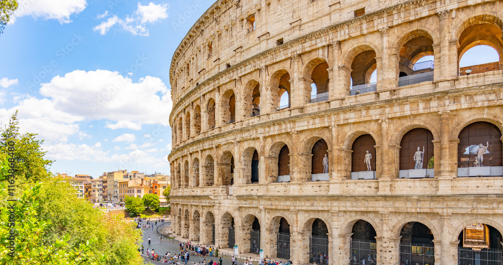 Obraz premium The majestic Colosseum stands prominently under a bright sky in Rome. Visitors admire its architecture, while trees and bustling city life create a vibrant atmosphere.