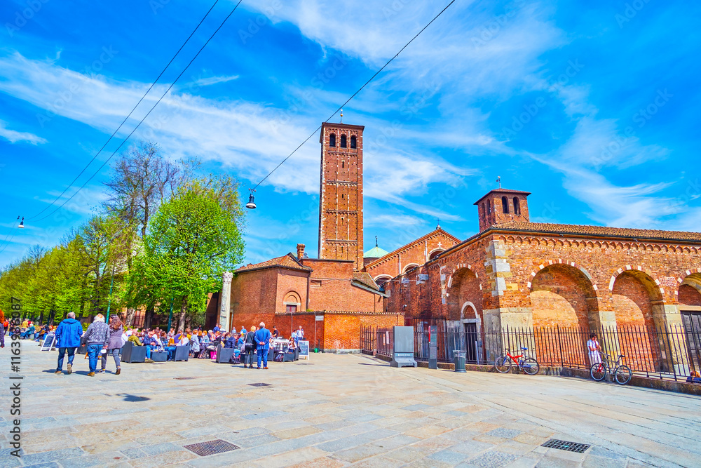 Outdoor restaurants on Piazza Sant'Ambrogio with towers of Basilica of Sant'Ambrogio on background, Milan, Italy