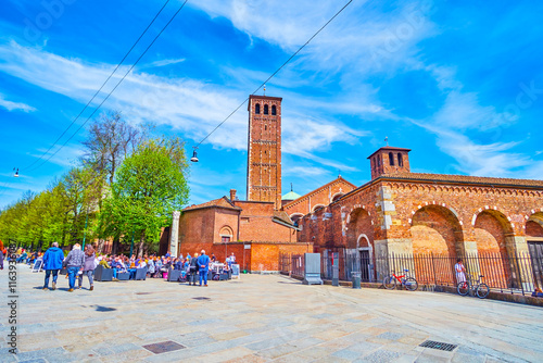 Outdoor restaurants on Piazza Sant'Ambrogio with towers of Basilica of Sant'Ambrogio on background, Milan, Italy