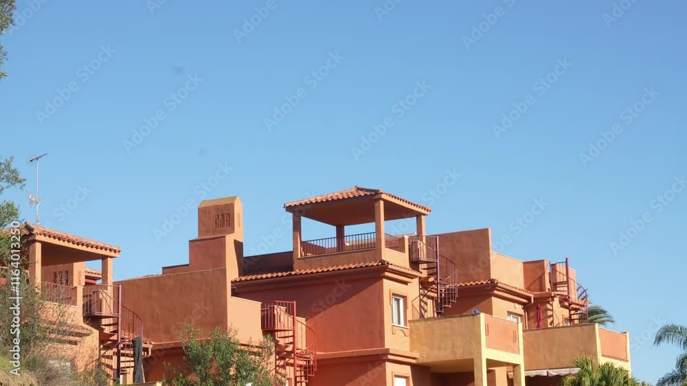 Cluster of Mediterranean-style terracotta-colored apartments featuring rooftop terraces and traditional architectural details like tiled roofs and open balconies. Set against a vibrant blue sky, the Cluster of Mediterranean-style terracotta-colored apartments featuring rooftop terraces and traditional architectural details like tiled roofs and open balconies. Set against a vibrant blue sky, the
