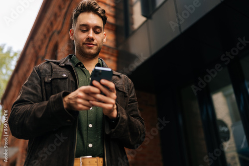 Low-angle view of handsome bearded man standing on city street, using smartphone to check messages, browse internet, stay connected enjoying urban environment. Concept of urban lifestyle.