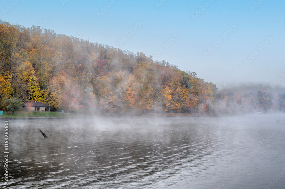 Mist drifts over a calm lake surrounded by a forest with trees displaying vibrant autumn colors. A small building is nestled among the trees, and the sky is clear blue above. Mist drifts over a calm lake surrounded by a forest with trees displaying vibrant autumn colors. A small building is nestled among the trees, and the sky is clear blue above.