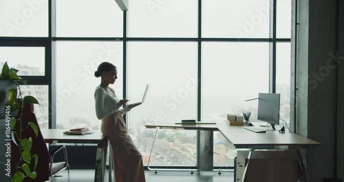 A young woman working on a laptop in a modern office with panoramic windows overlooking an urban coastal view, symbolizing productivity, creativity, and contemporary work culture