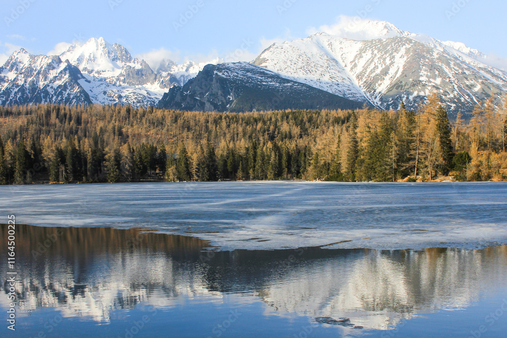 High in the mountains, the ice on a mountain lake melts in spring. Reflections of the mountains and sky are visible in the water. High in the mountains, the ice on a mountain lake melts in spring. Reflections of the mountains and sky are visible in the water.